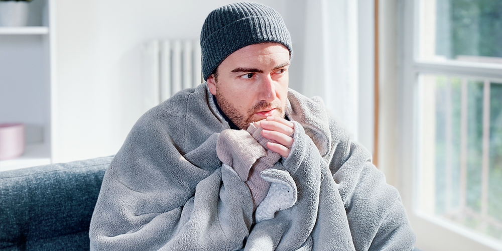 Male homeowner sits bundled up on couch freezing thanks to a furnace blowing cold air.