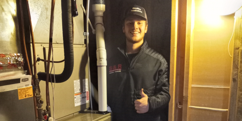 S&R technician in hat and coat performs annual furnace maintenance in customer's home.